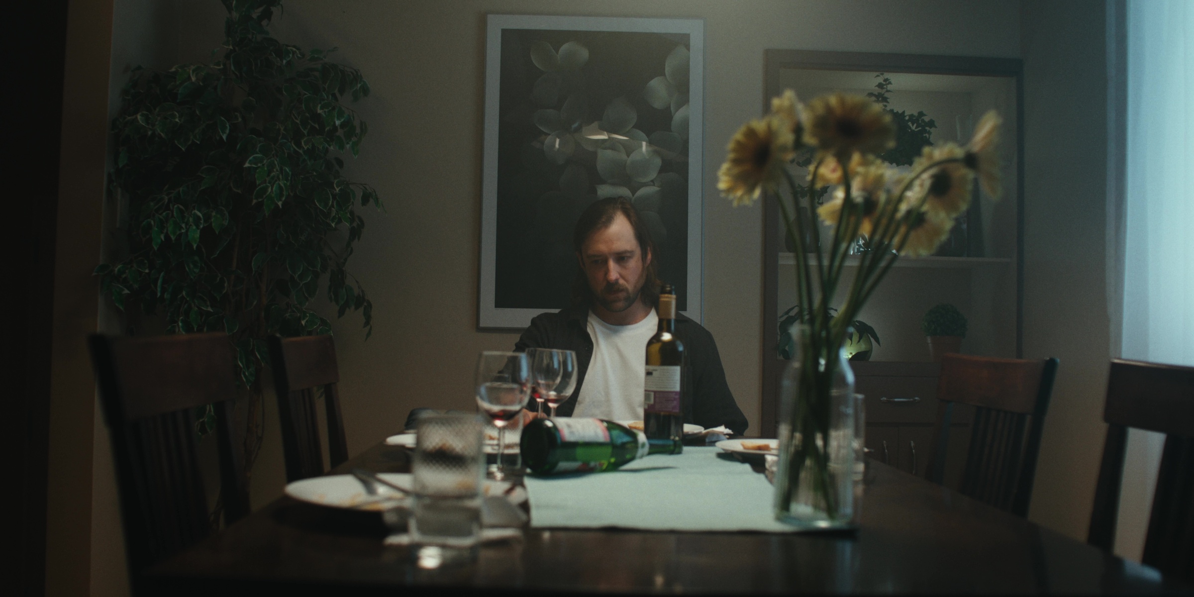 A man sits alone at a dining table beside dried flowers — Bottom of the bottle