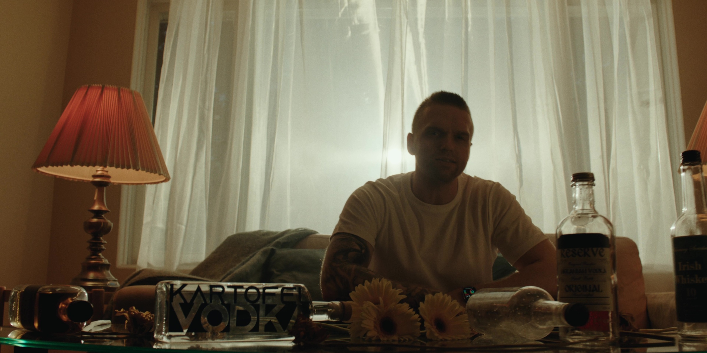A man sits behind a table of flowers and bottles near a bright window — Bottom of the bottle