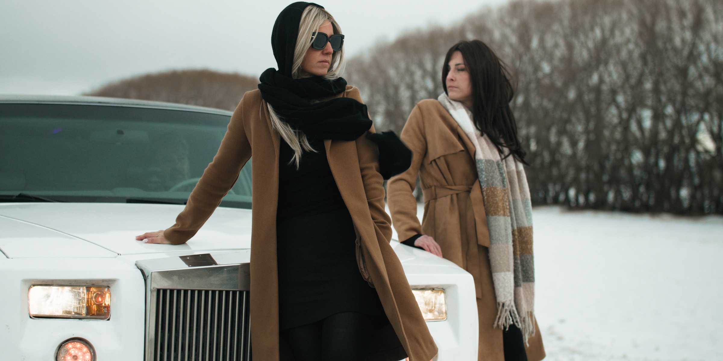 Two women in camel coats stand beside a white car in the snow — Chiquel