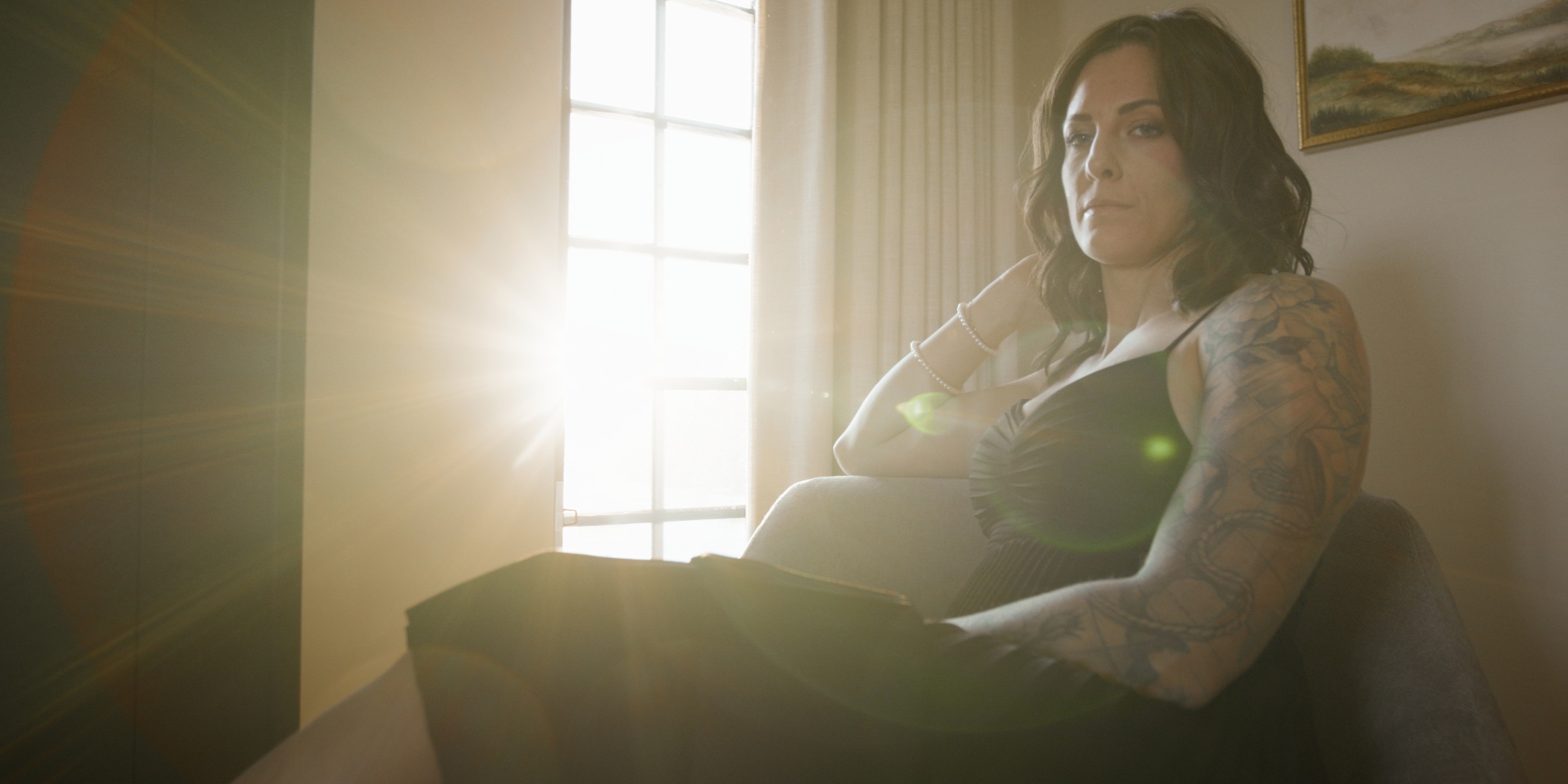 Sunlight falls across a tattooed woman seated beside a window — Chiquel