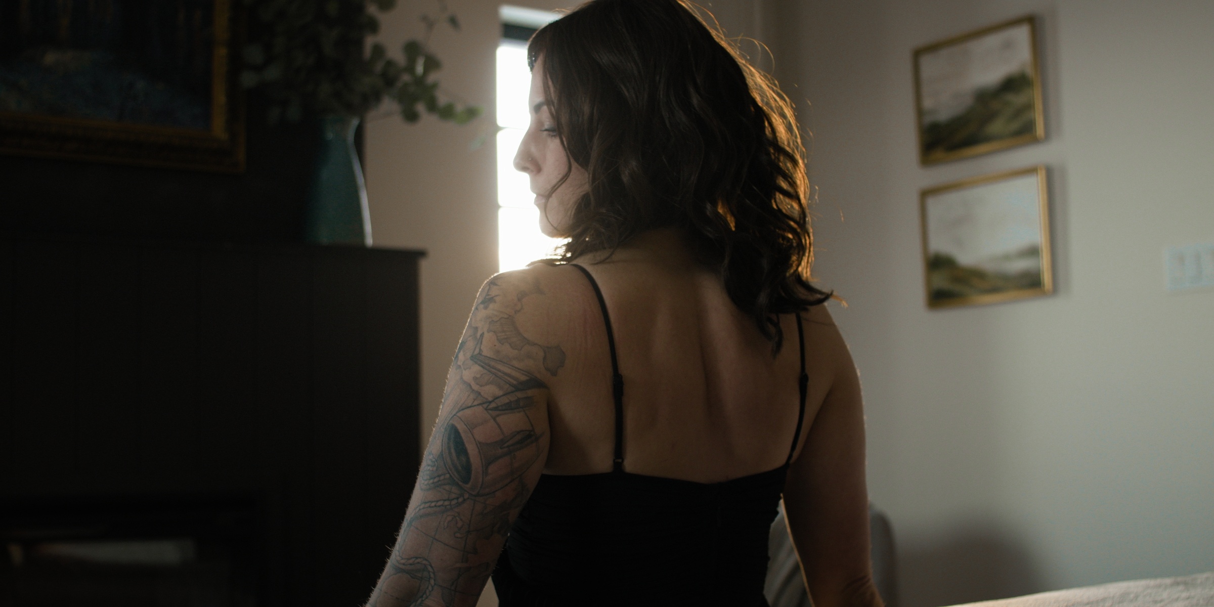 A tattooed woman turns beside a piano in warm window light — Chiquel