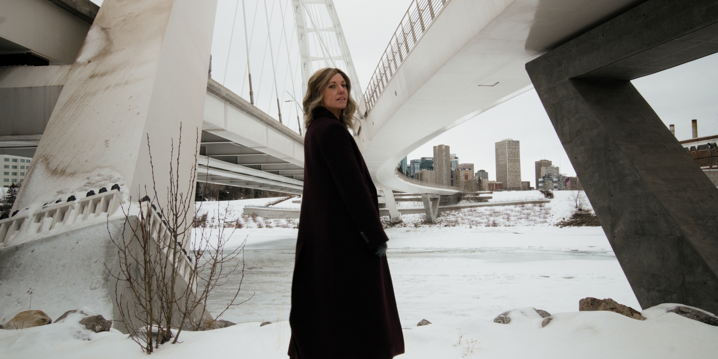 A woman stands in the snow beneath a sweeping concrete bridge — Chiquel