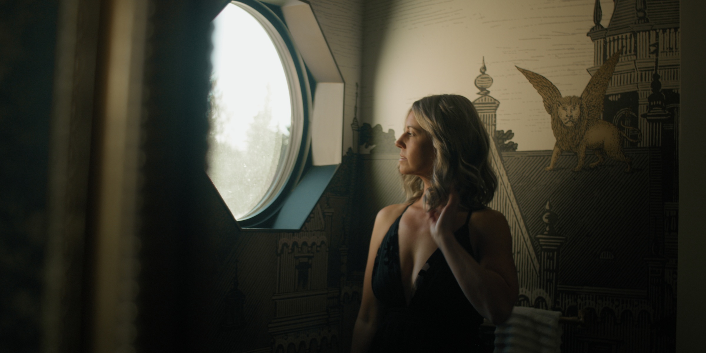 A woman stands beside a round window in a dim room — Chiquel