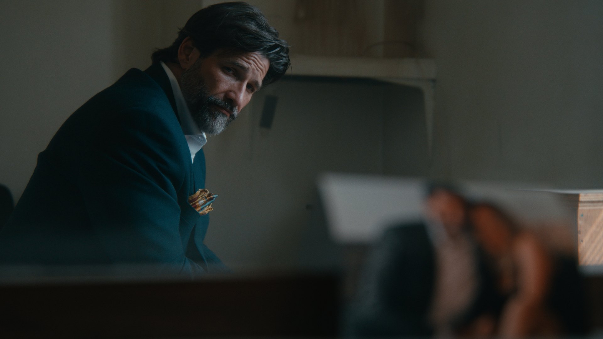 A bearded man in a dark suit seated in a dim interior — Crossroads