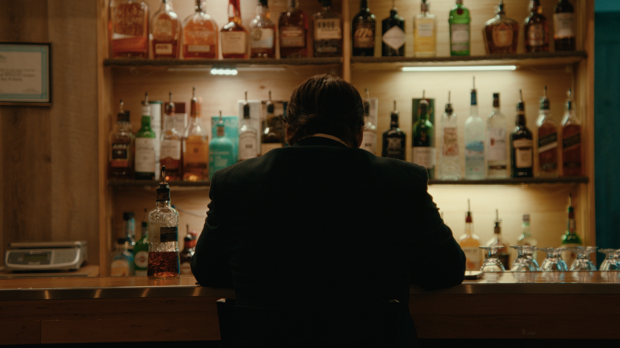 A man sits at a bar facing shelves of liquor bottles — Crossroads
