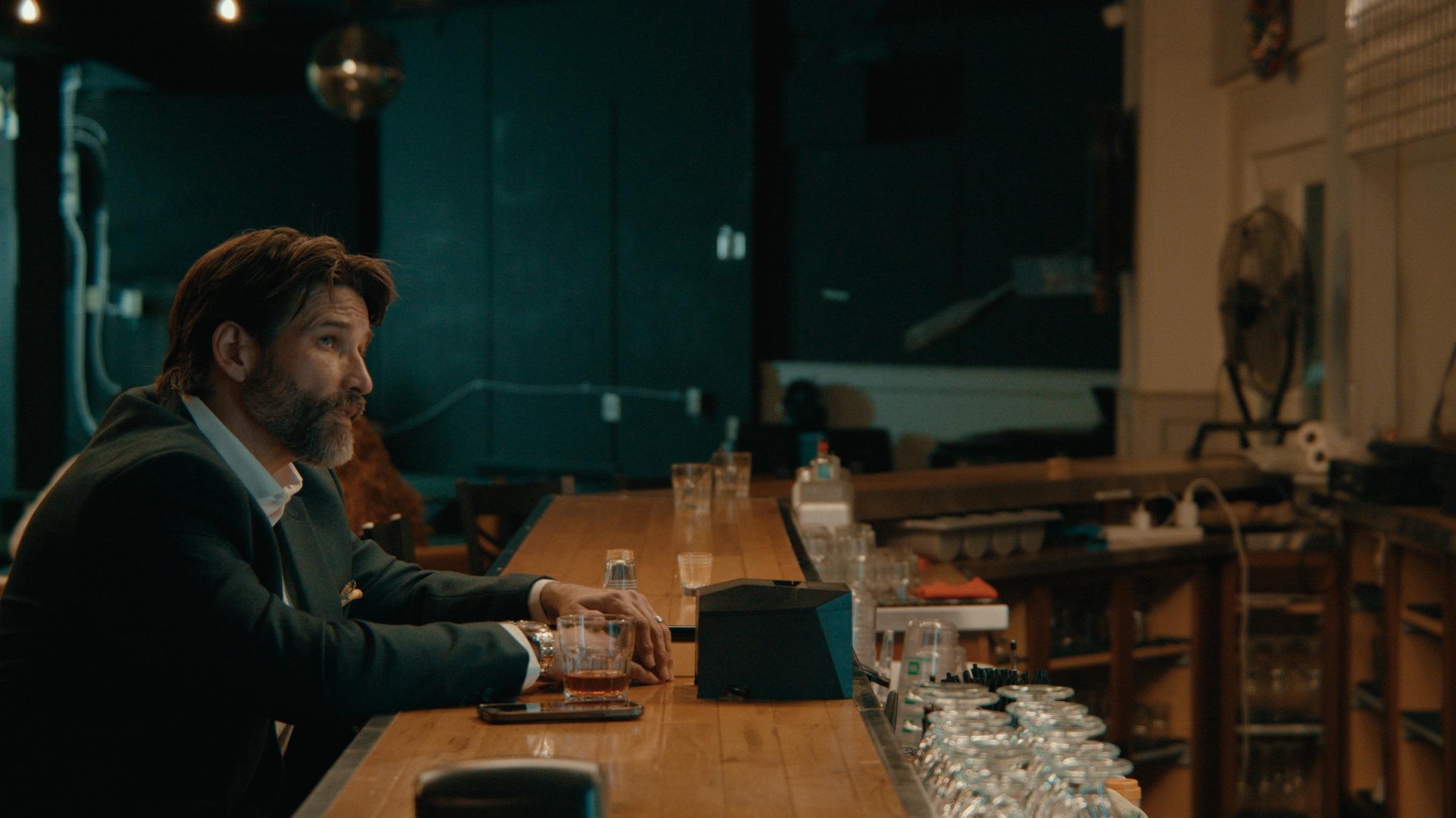 A man in a dark suit talks to a long-bearded bartender — Crossroads