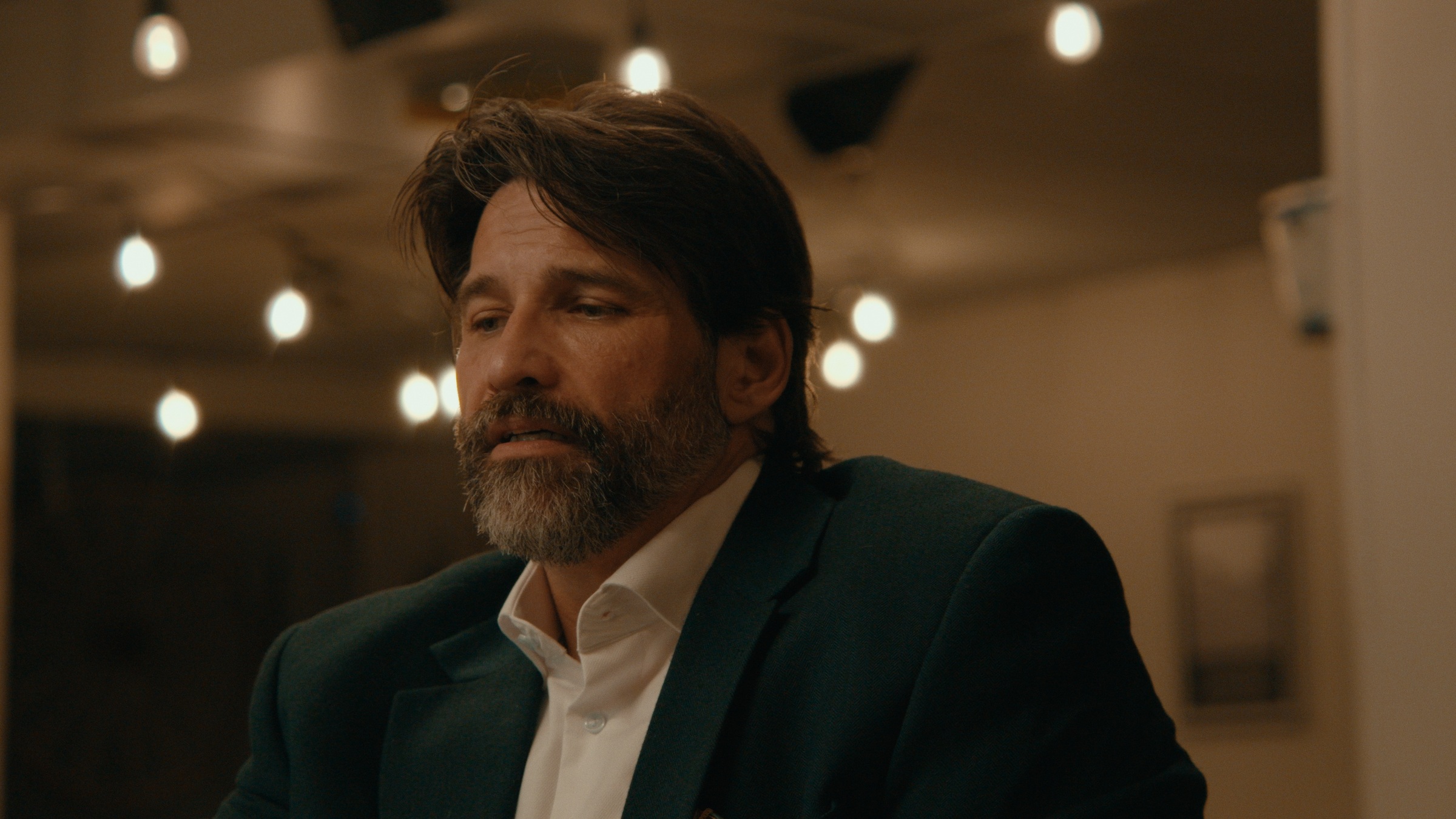A bearded man sits beneath warm hanging lights at a bar — Crossroads