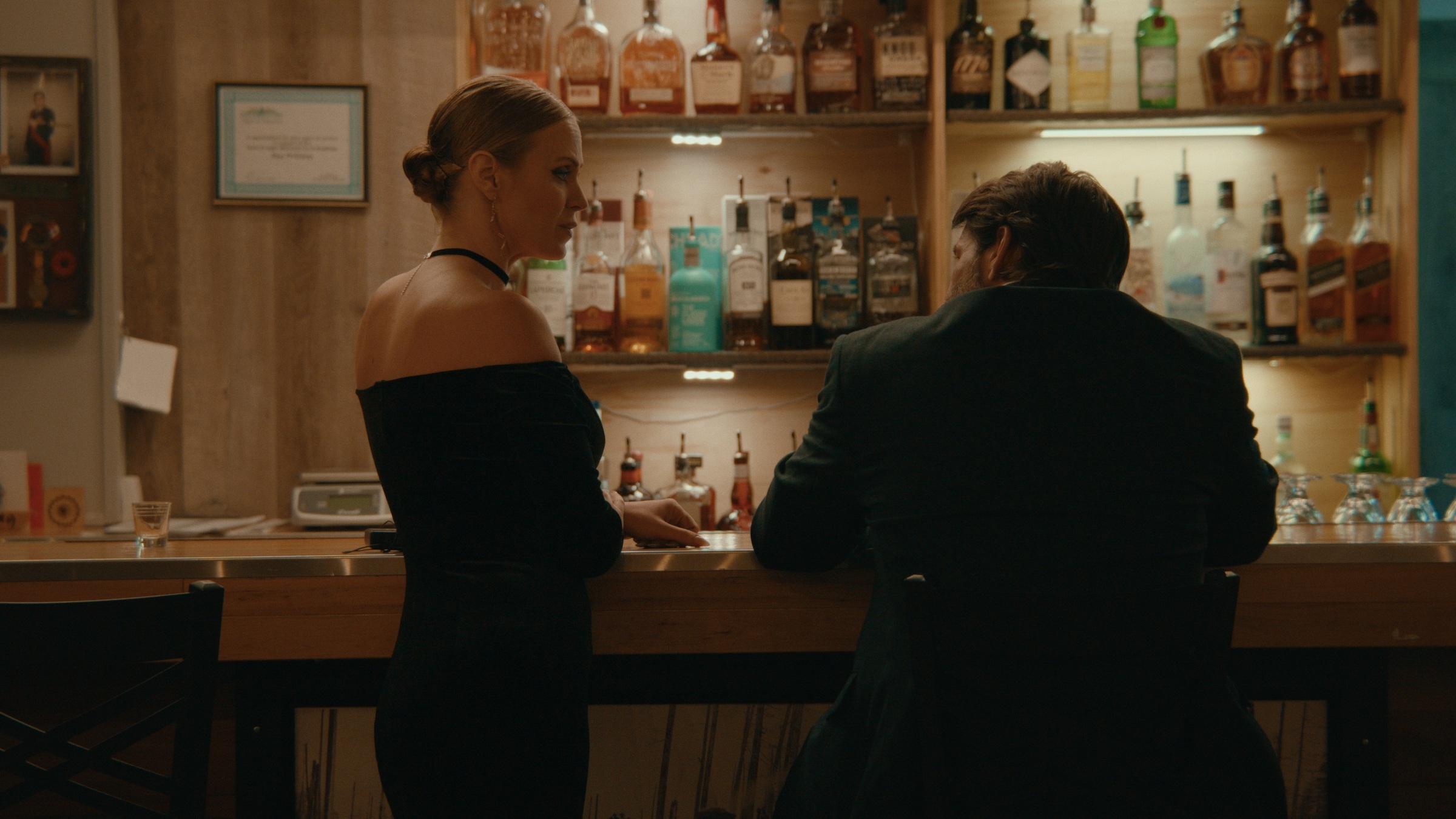 A woman and a man sit together facing a warmly lit bar — Crossroads