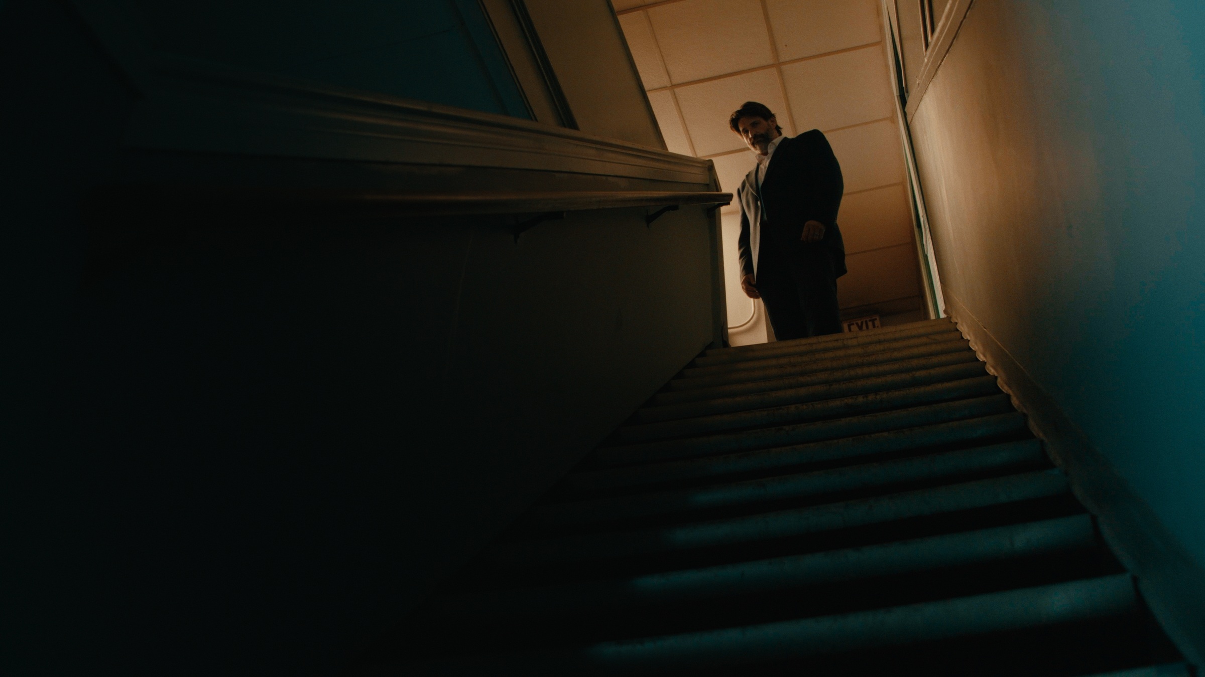 A man stands at the top of a dim stairwell above teal steps — Crossroads