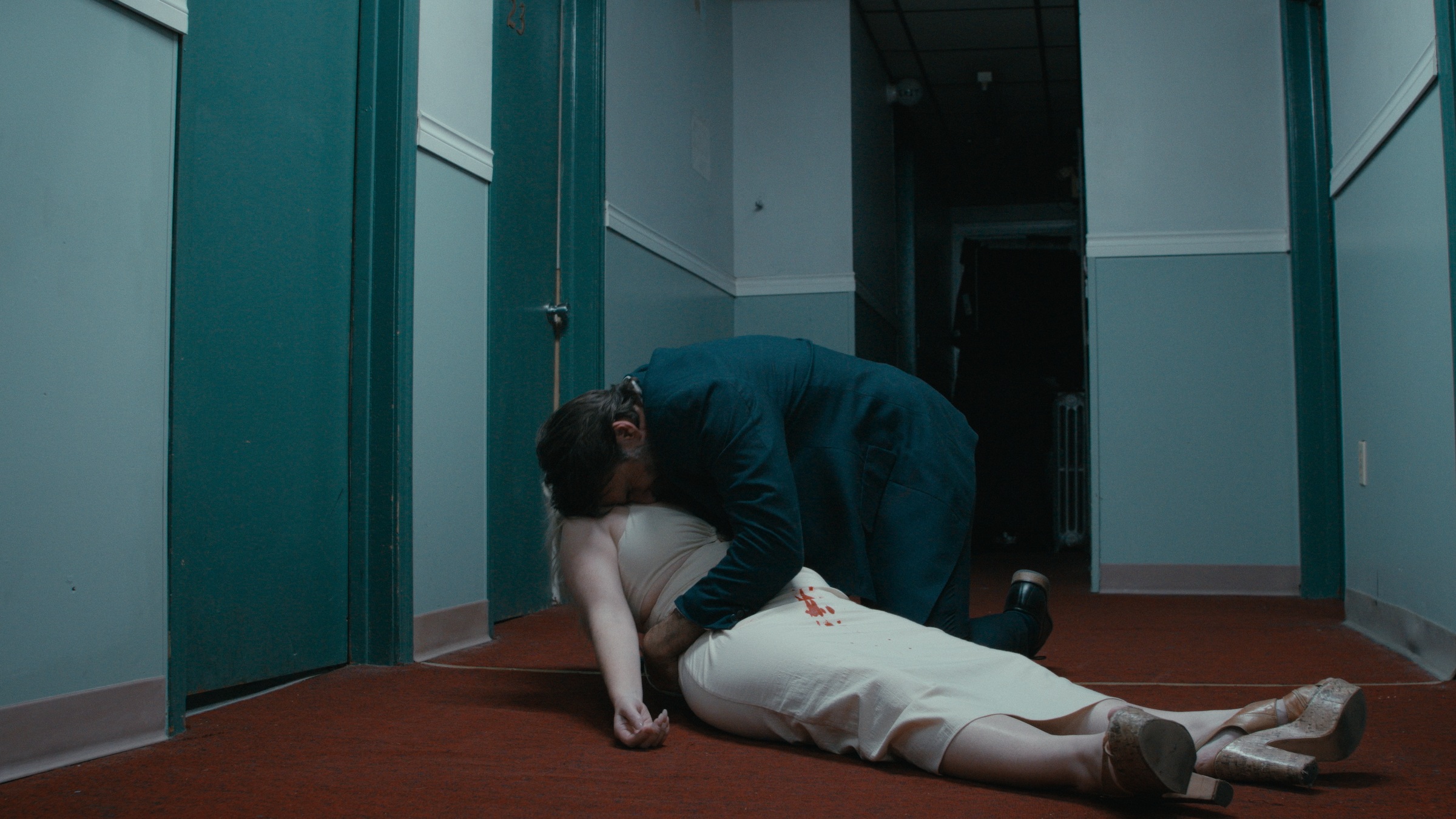 A man kneels beside a woman lying on a red-carpeted hallway floor — Crossroads