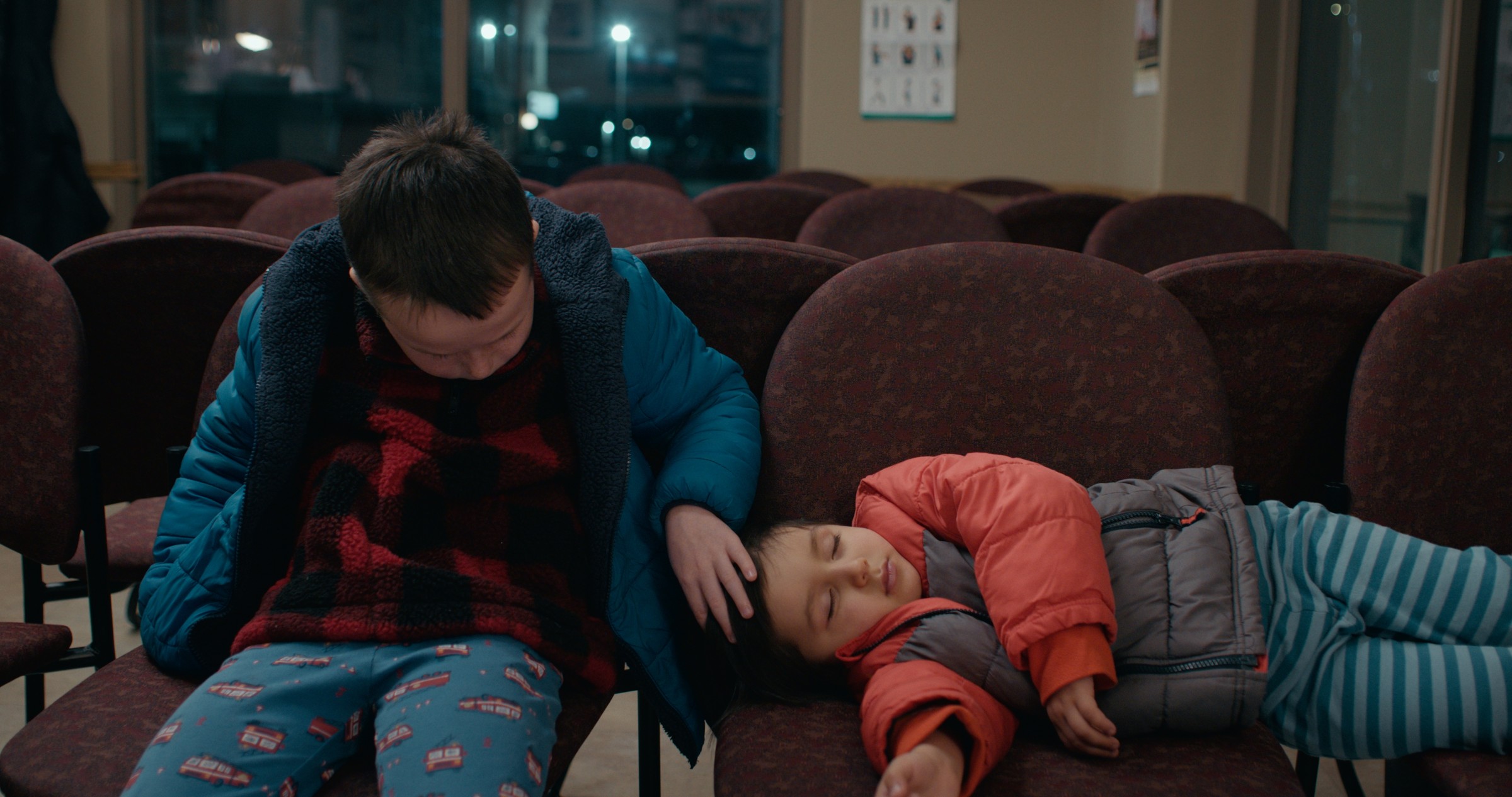 A boy in a blue coat sits beside a reclining child in a waiting room — Emergency