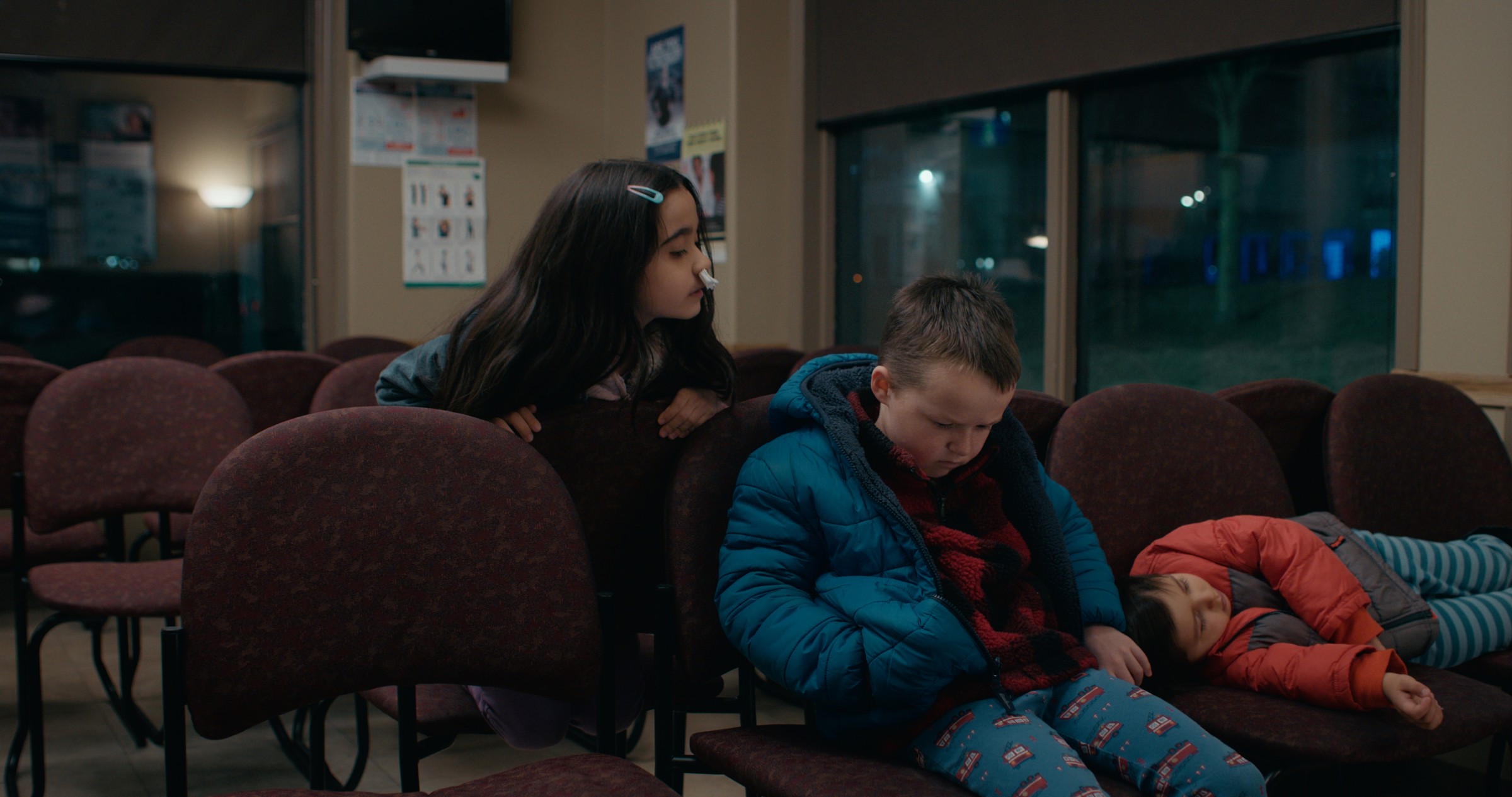 Children sit among rows of burgundy waiting-room chairs at night — Emergency