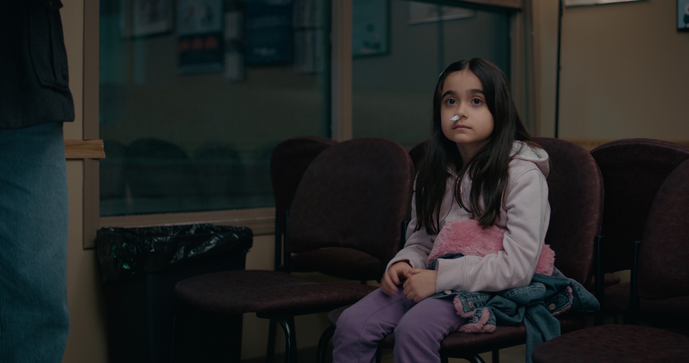 A girl in a pink coat sits alone among empty waiting-room chairs — Emergency