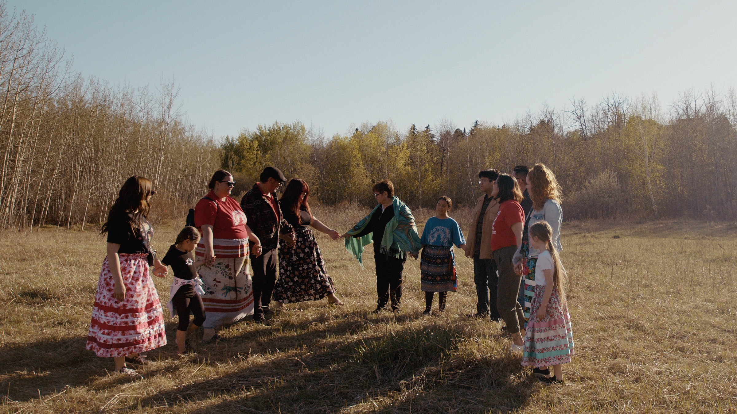 A group holds hands in a circle in a grassy clearing — Home Fire