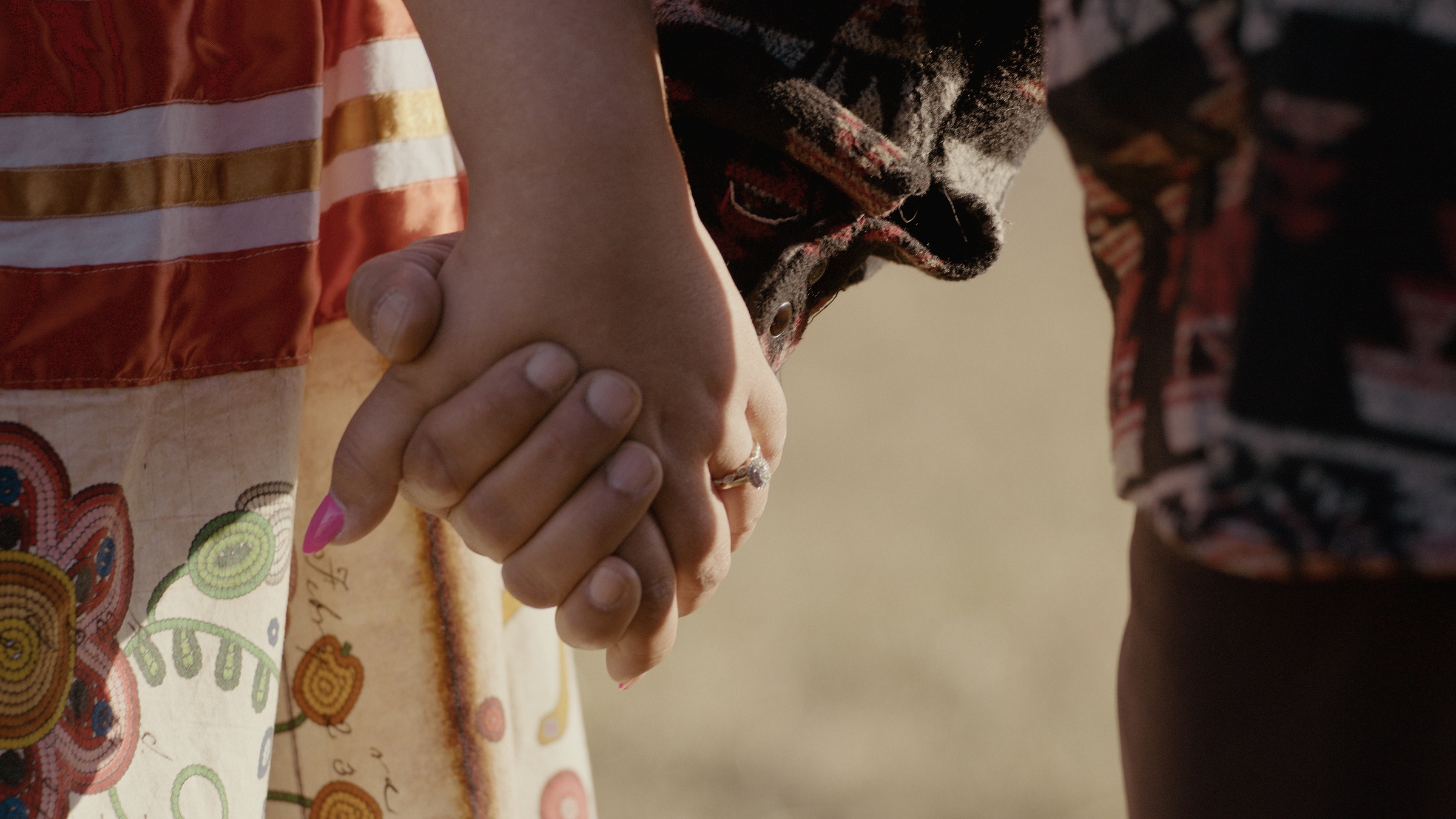 Close-up of two people holding hands beside colorful skirts — Home Fire