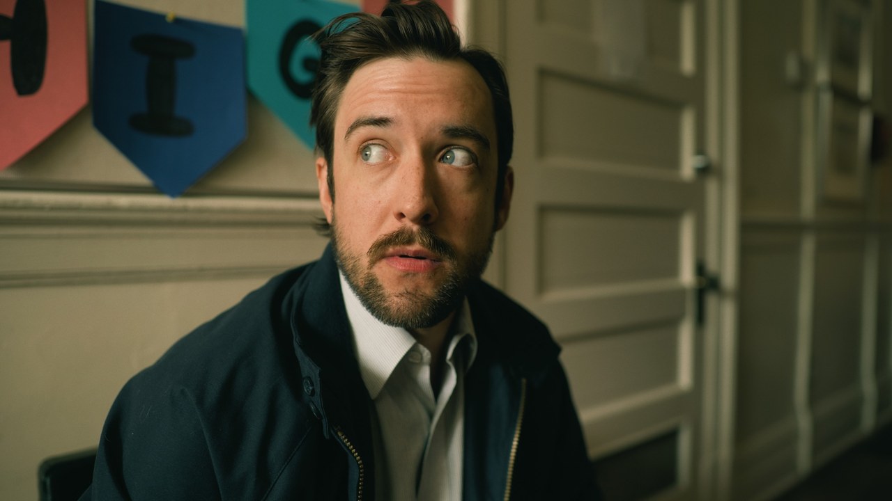 A man looks to the side while seated in a school hallway — Little Fangs