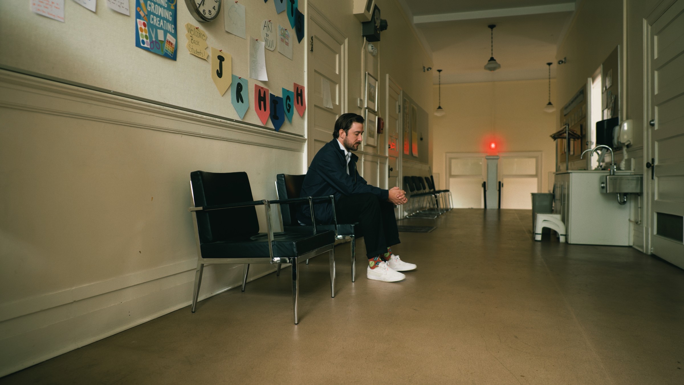 A man sits on a row of chairs in a school hallway — Little Fangs