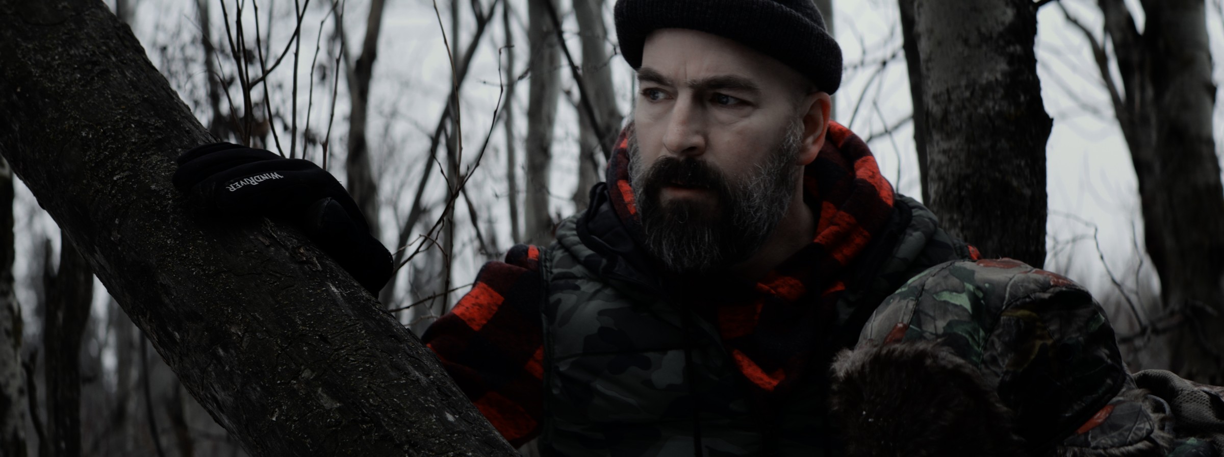 A bearded man in a cap stands beside a tree in a forest — Markings of Murder