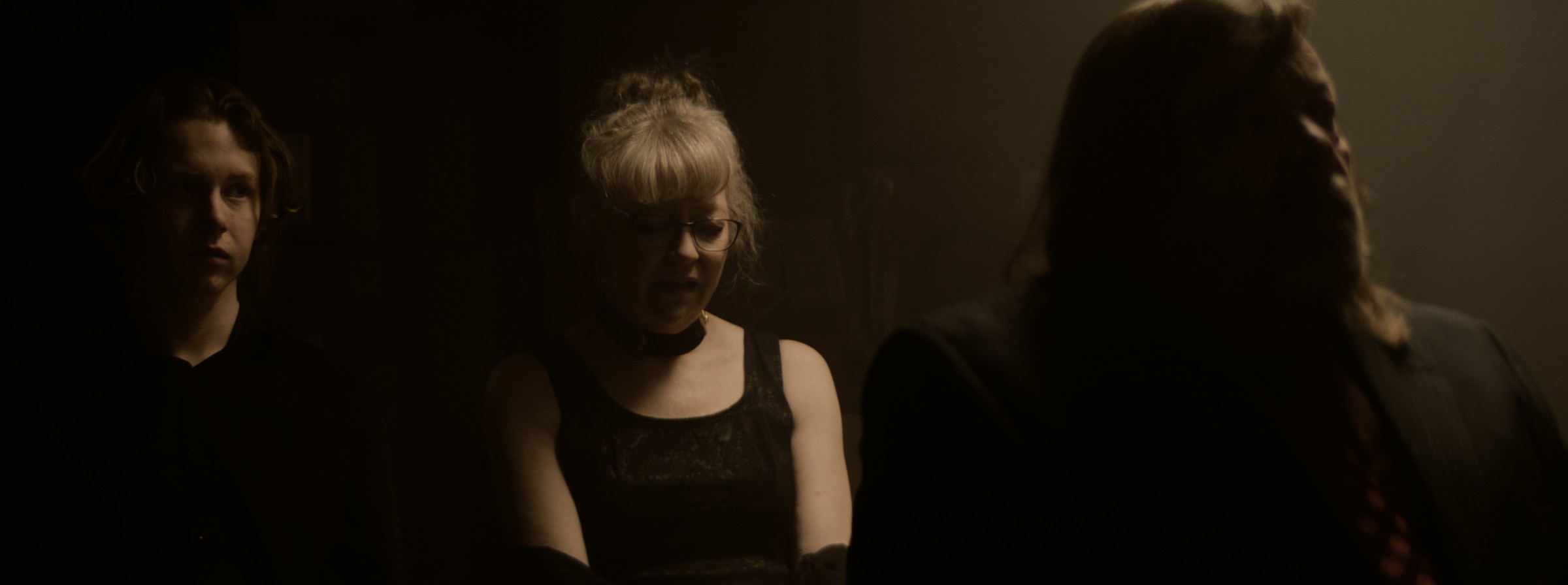 Three people stand together in a dim interior — Markings of Murder