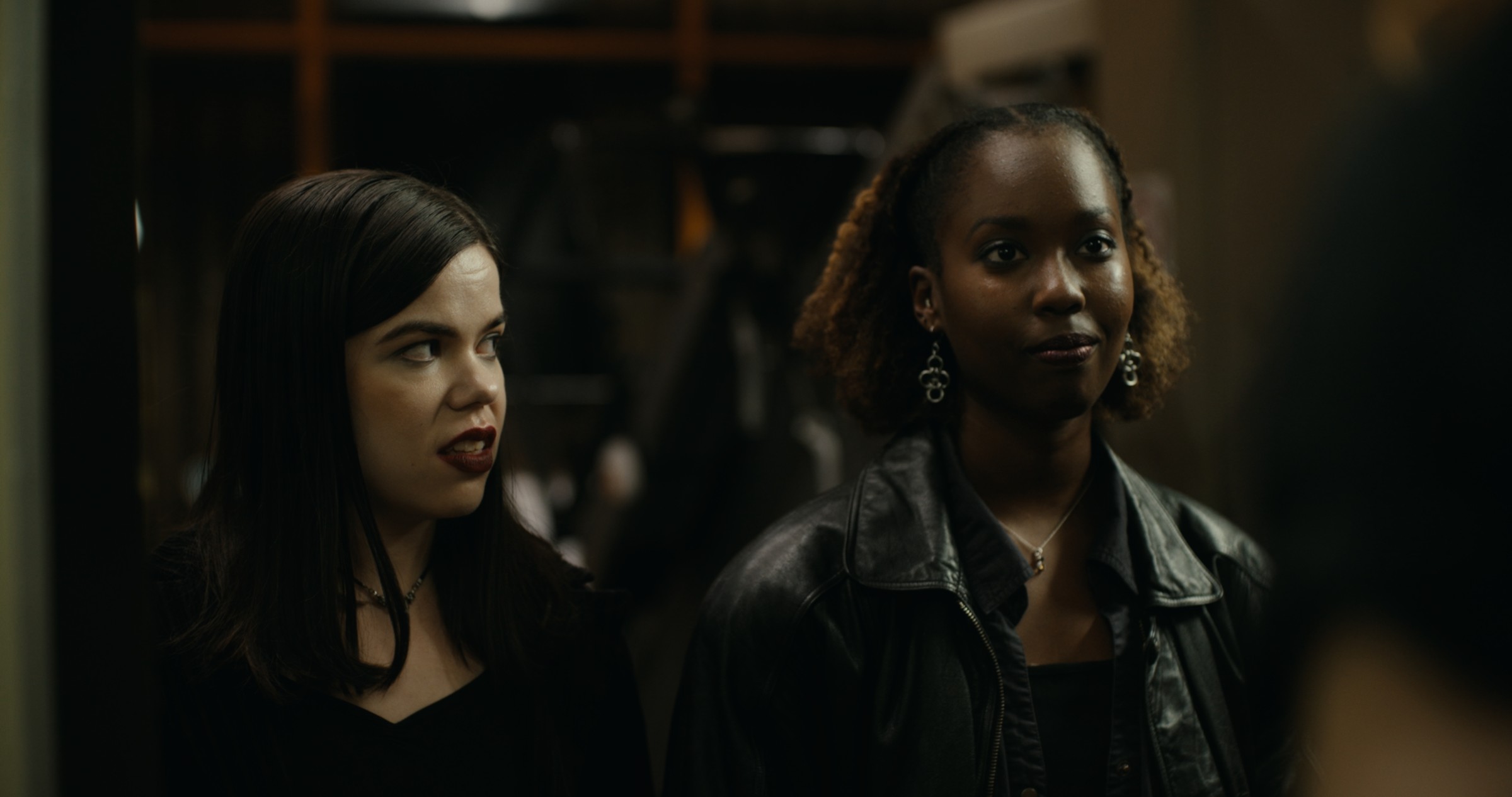 Two women stand together in a dim room — Nemesis