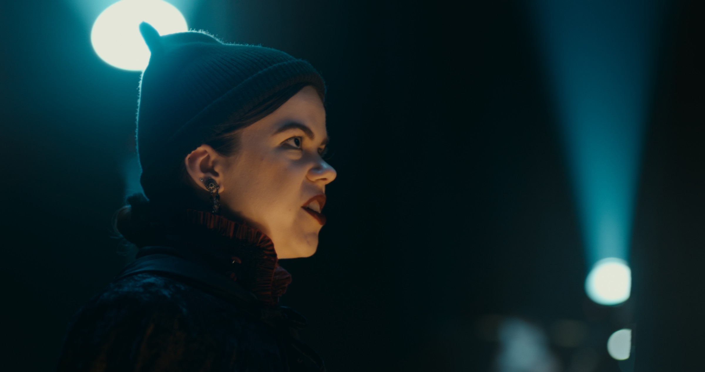 A woman in a black headband looks sideways beneath a cool light — Nemesis