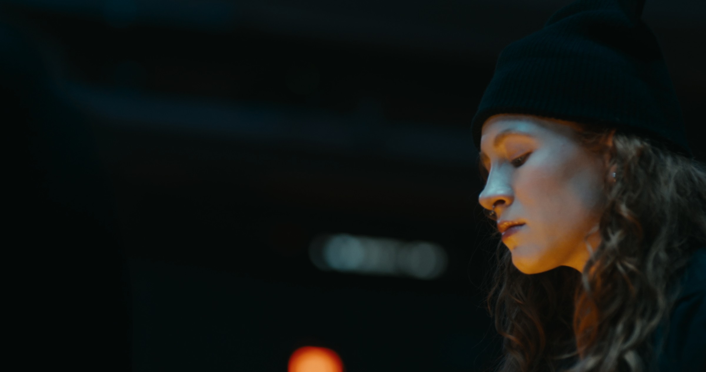 A woman's face is lit by warm light against a dark background — Nemesis