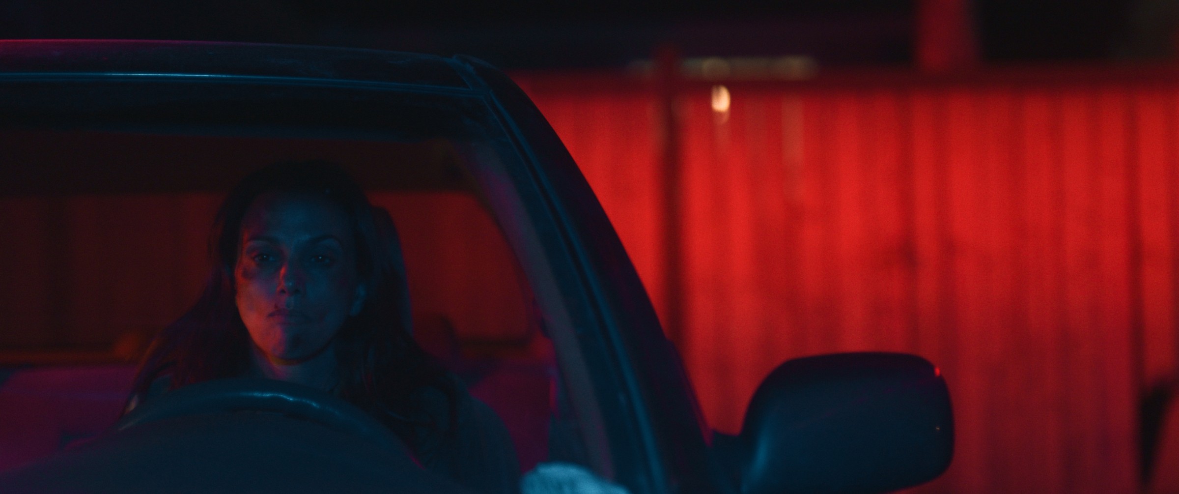 A woman sits in a car beside a red-lit wall at night — The Table Between Us