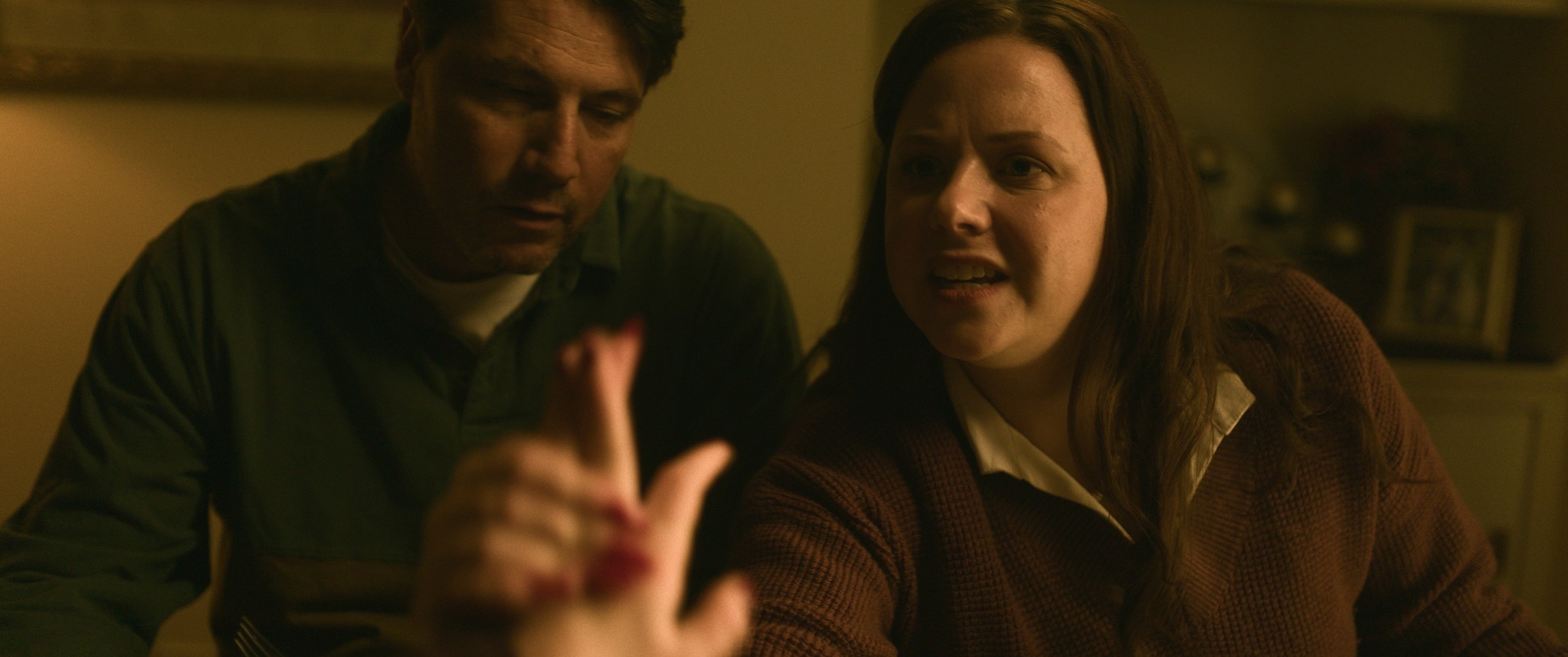 A man and woman talk together in a warmly lit interior — The Table Between Us
