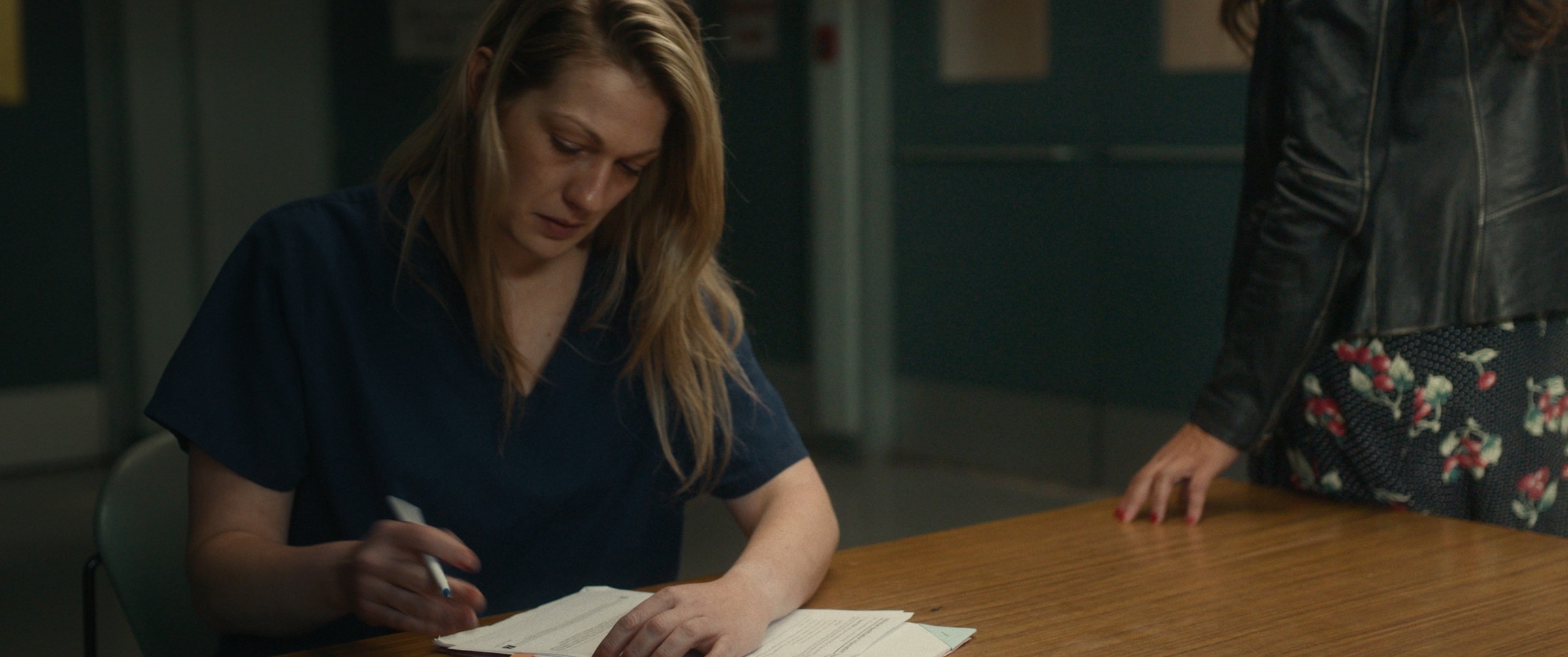 A woman in blue writes on papers spread across a table — The Table Between Us