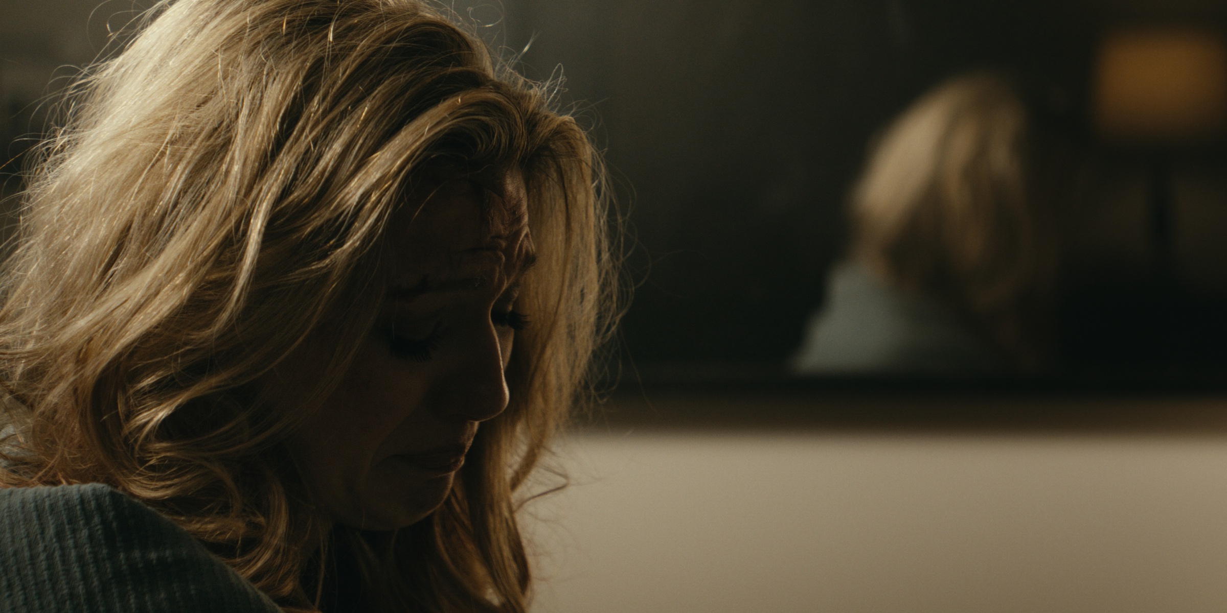 A woman looks down beside a mirror in a dimly lit room — Trinkets
