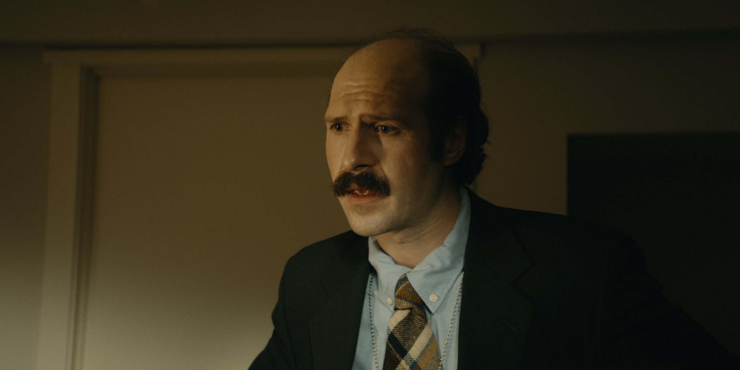 A mustached man in a suit and patterned tie stands in a softly lit room — Trinkets