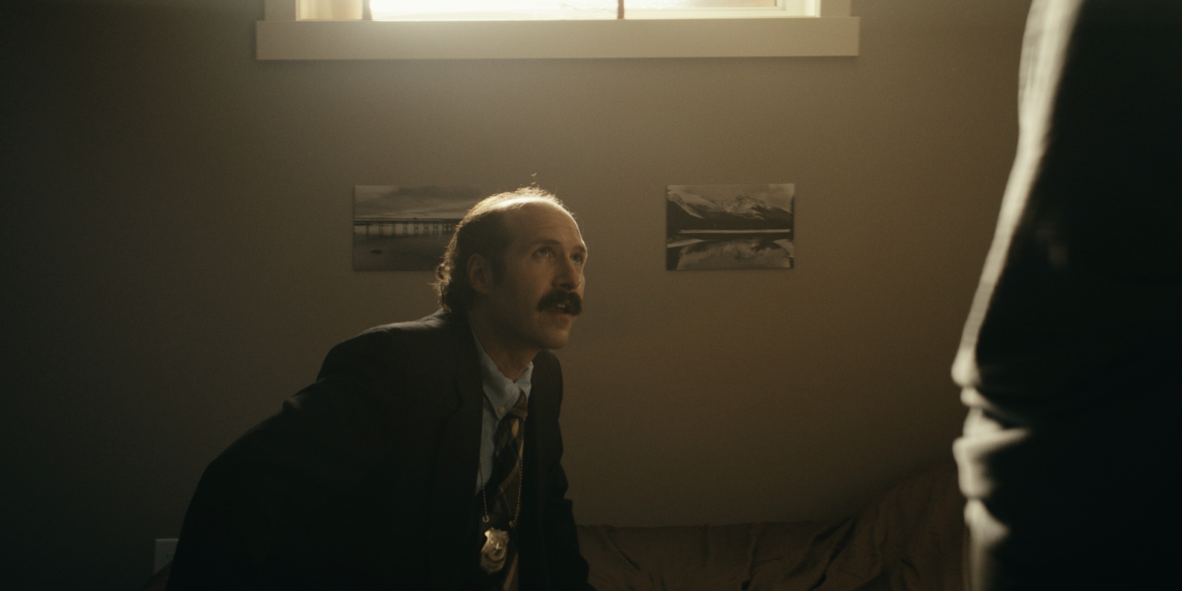 A seated man looks up toward a woman beside a bed — Trinkets