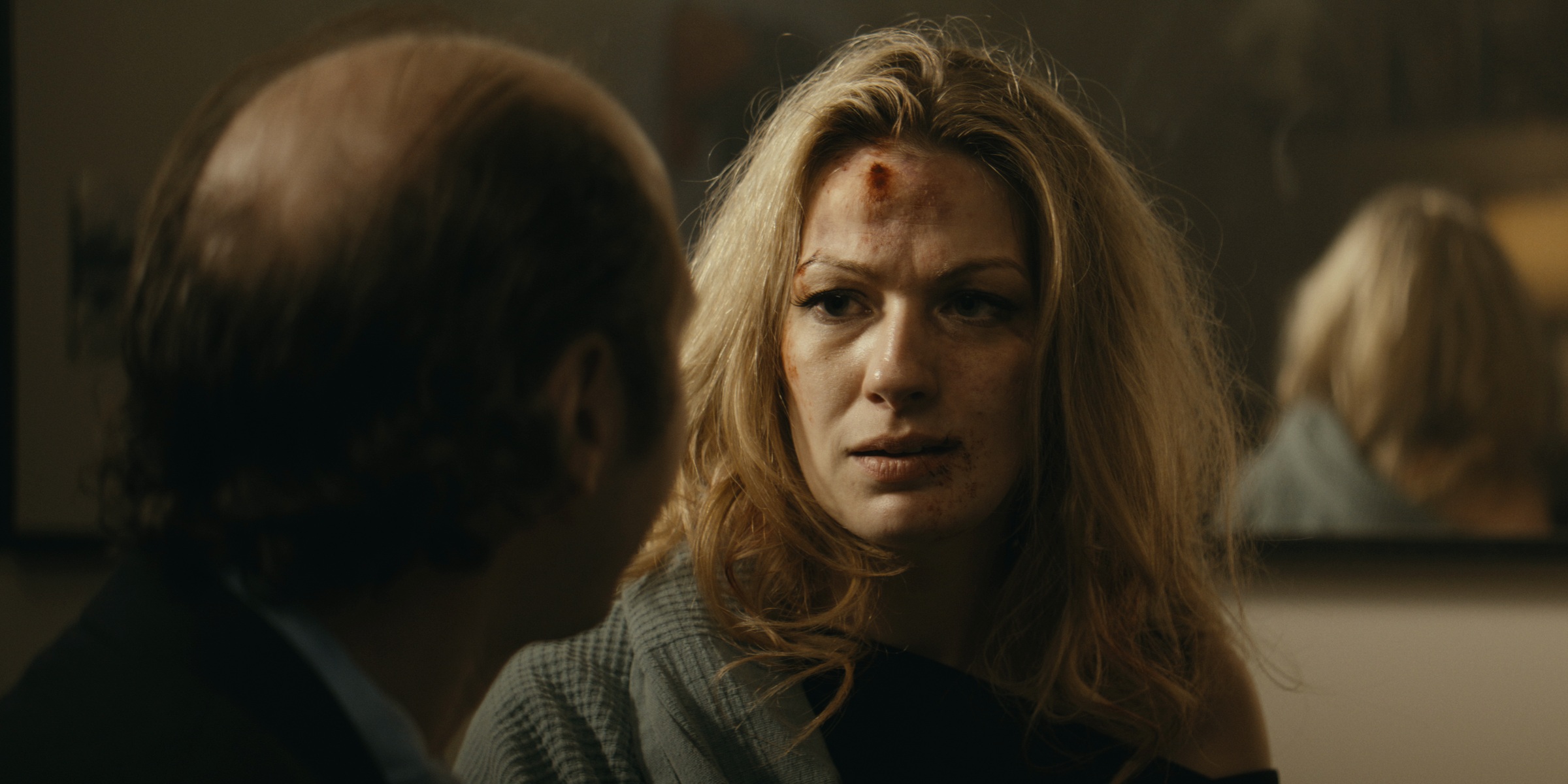 A woman with blood on her forehead looks toward a man in a dim bedroom — Trinkets