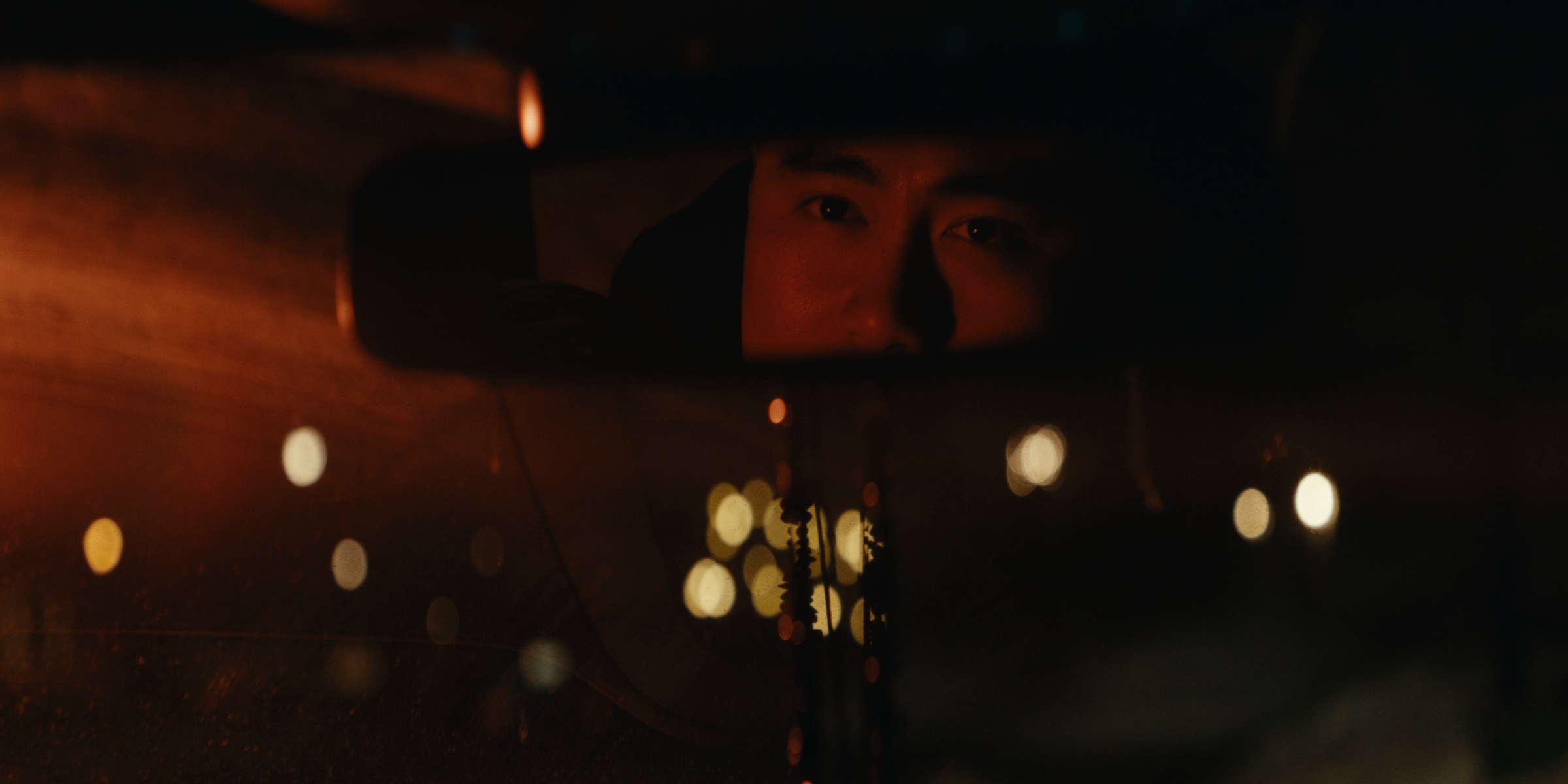 A driver reflected in a rearview mirror amid warm night lights — Trinkets