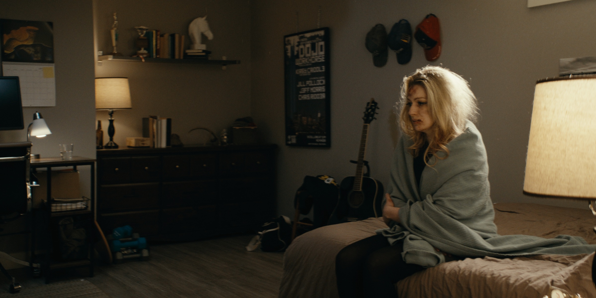 A woman wrapped in a blanket sits on a bed beside a lamp and guitar — Trinkets