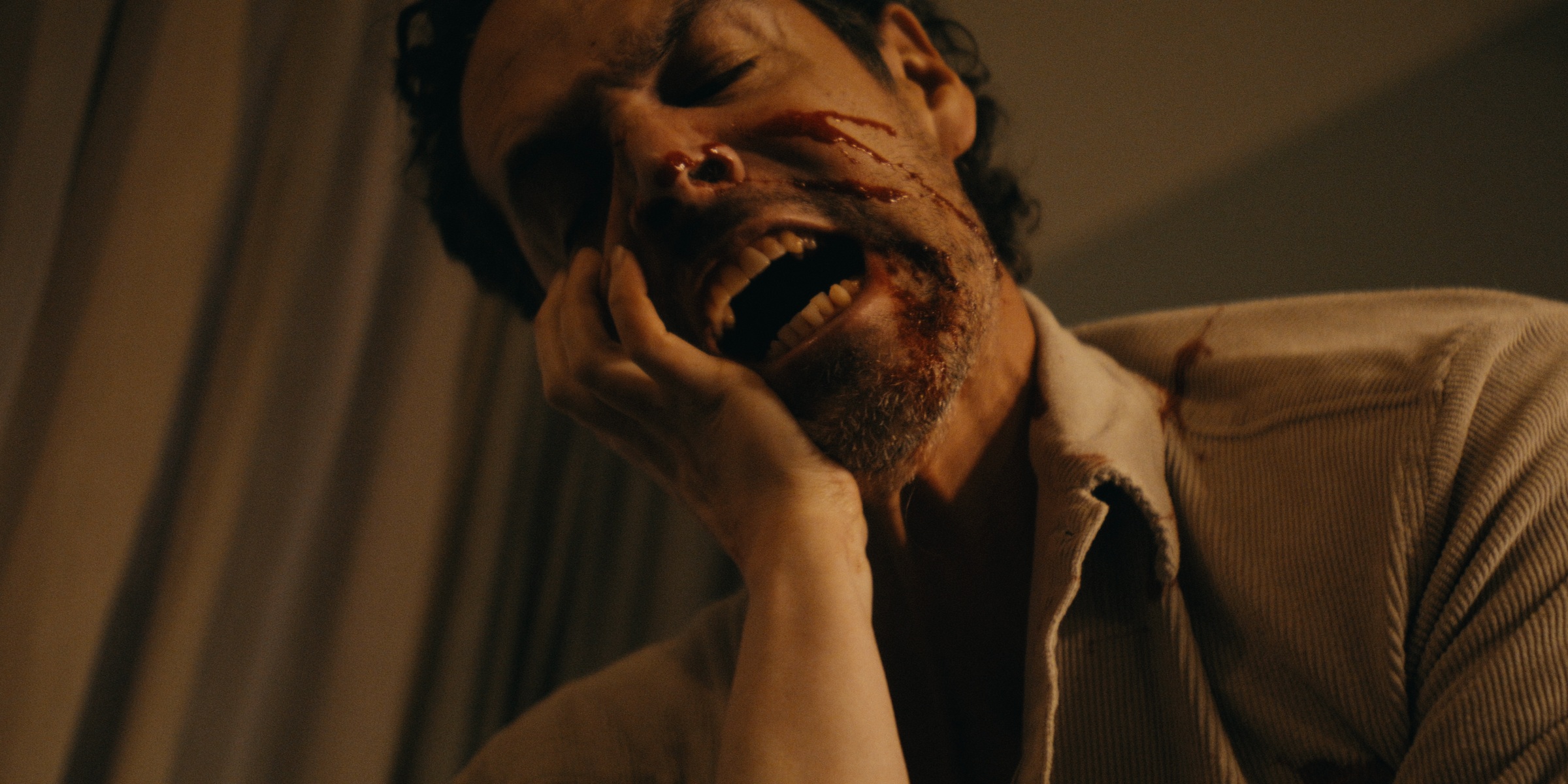 A man with blood on his face holds his cheek in a warm, dim interior — Trinkets